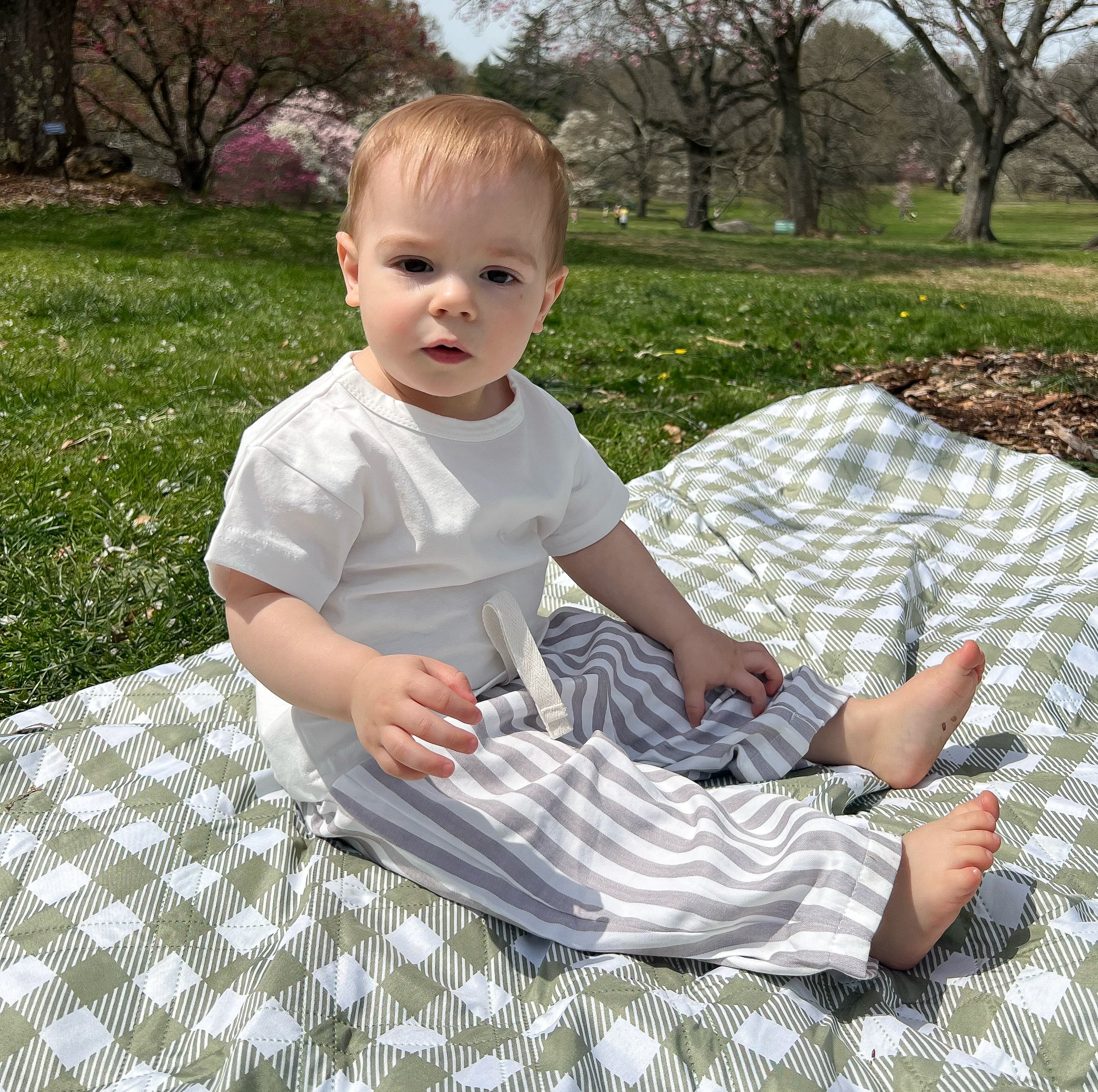 A baby sitting on a checkered picnic blanket wearing a white t-shirt and white and gray striped pants with a relaxed fit.