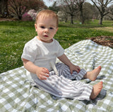 A baby sitting on a checkered picnic blanket wearing a white t-shirt and white and gray striped pants with a relaxed fit.