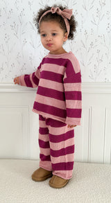 Toddler wearing a striped pink and purple outfit with a bow in hair, standing against a white wall.