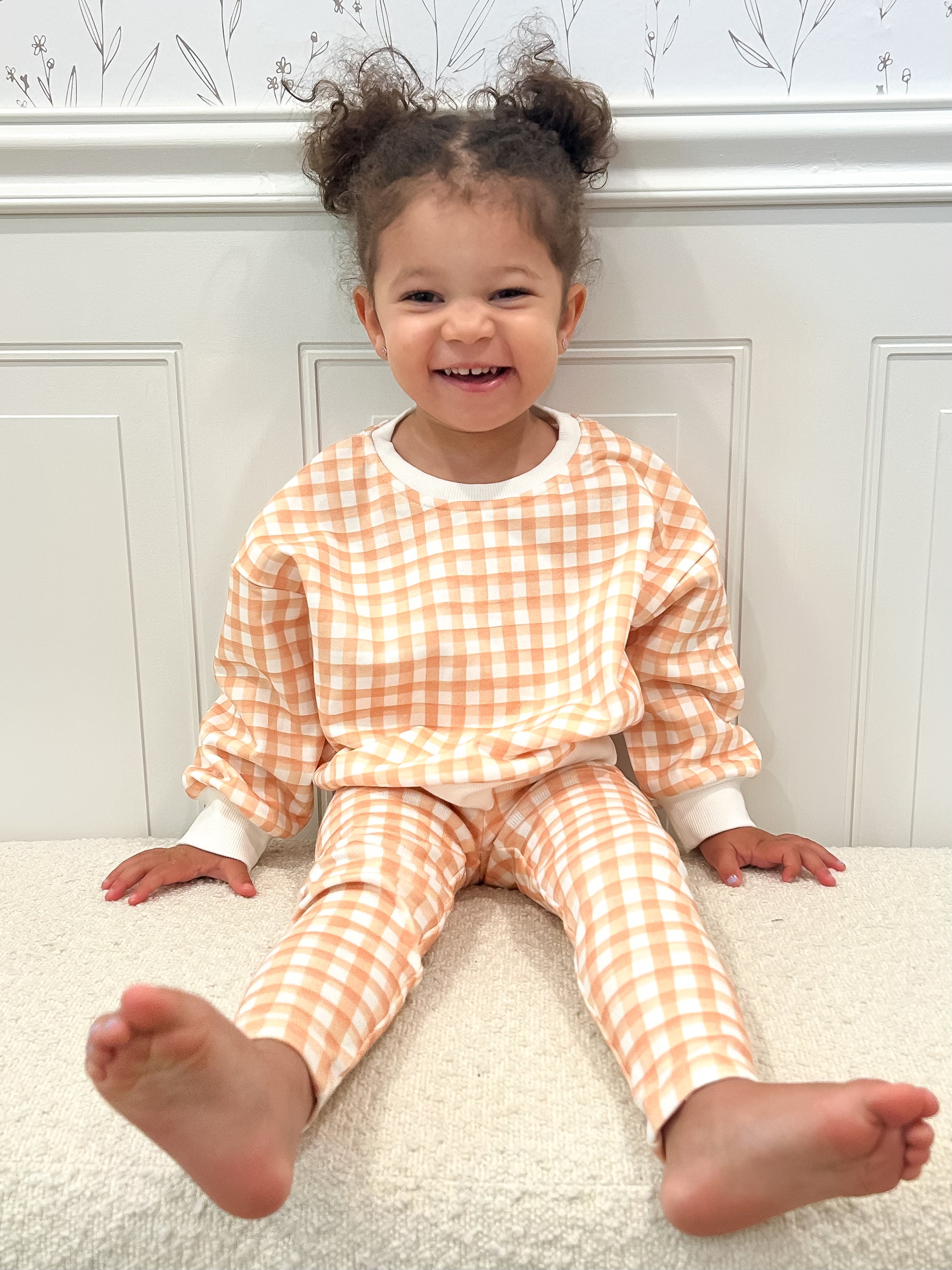 Toddler wearing an orange and white checkered outfit sitting on a sherpa ottoman.