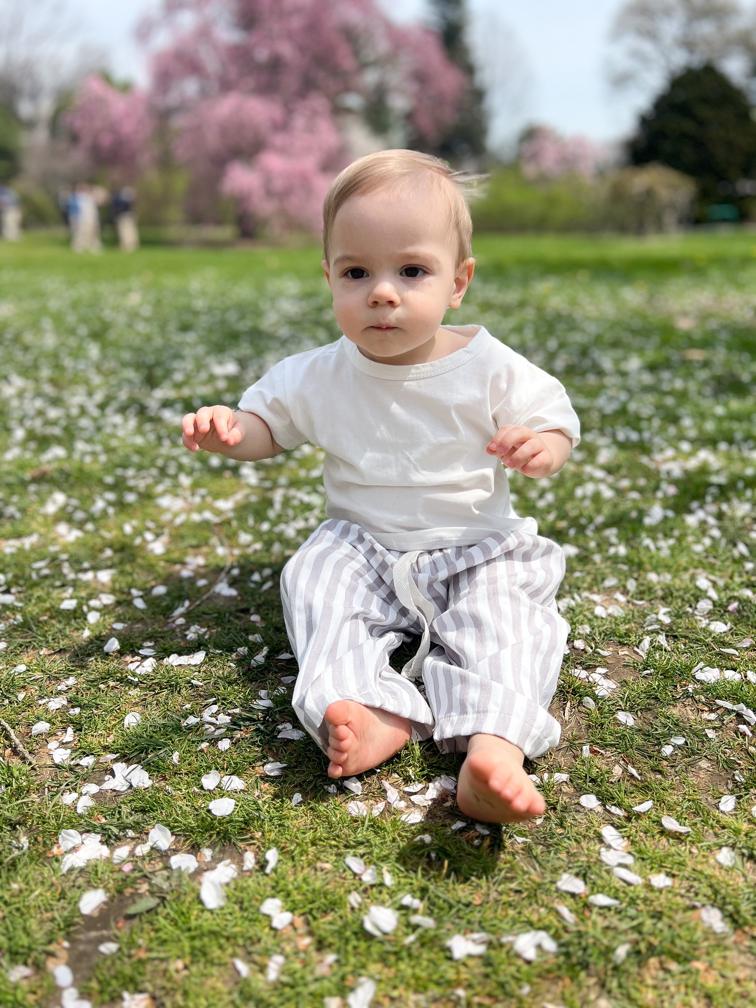 A baby sitting on grass surrounded by pink flower petals, wearing a white t-shirt and striped pants.