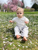 A baby sitting on grass surrounded by pink flower petals, wearing a white t-shirt and striped pants.