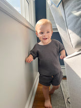 Toddler standing in a room wearing a charcoal waffle set with a window and gray sofa.