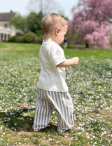 A toddler walking on grass with striped pants, with a tree in bloom in the background.