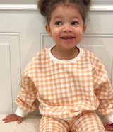 Toddler wearing an orange and white checkered outfit sitting on a beige sherpa ottoman.