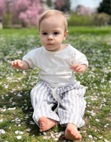 A baby sitting on grass with petals around, wearing a white t-shirt and striped pants.