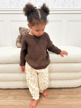 Child wearing a brown sweater and beige floral pants with a teddy bear on a sherpa ottoman.