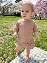 A baby standing on a checkered picnic blanket wearing a beige waffle short sleeve top and matching shorts in a field with cherry blossoms in the background.