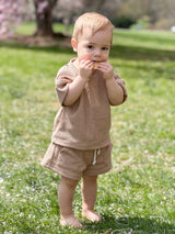 A baby eating a cracker standing barefoot on the grass wearing a tan waffle shorts sleeve and shorts outfit set with cherry blossom in the background.