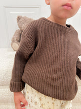 Child wearing a brown knitted sweater with a teddy bear in the background