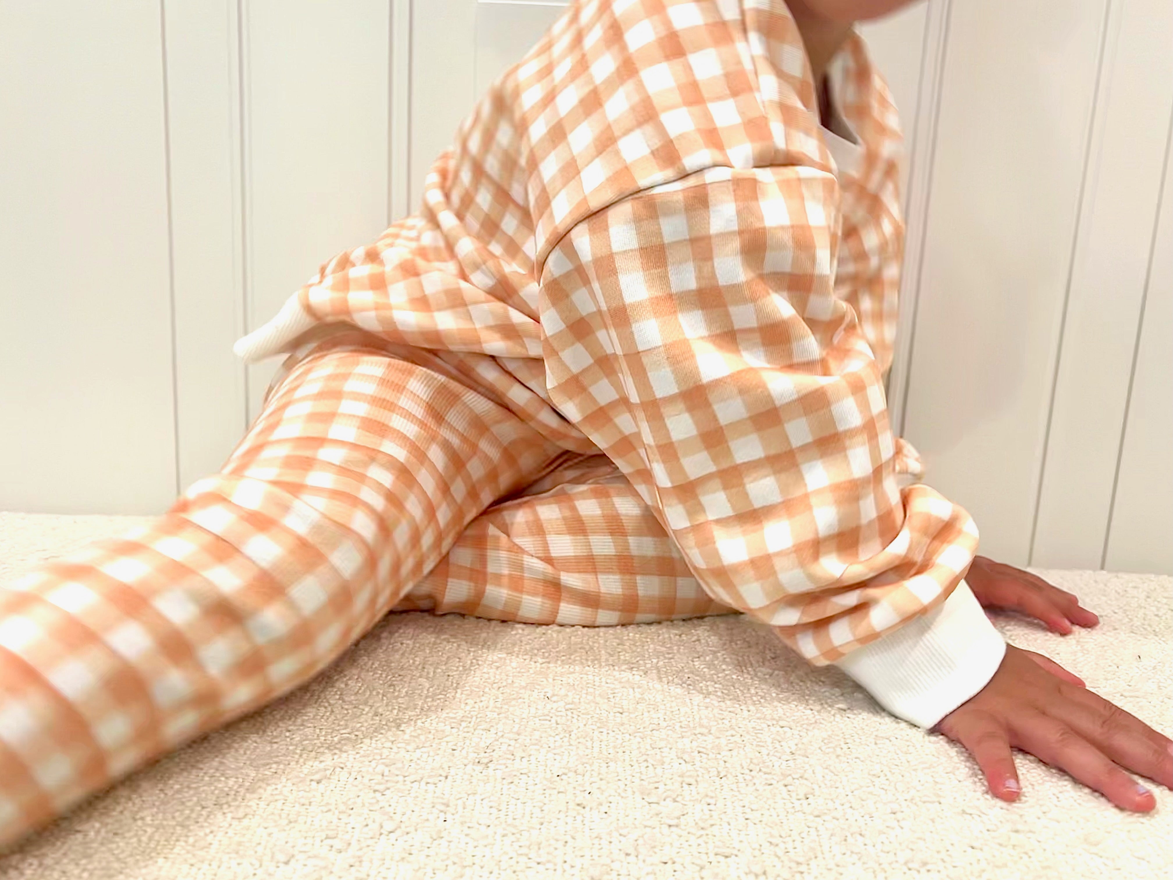 Toddler wearing orange and white checkered outfit set sitting on a sherpa ottoman leaning forward.
