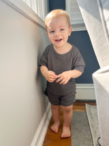 Toddler standing in a room wearing a charcoal outfit set with white and blue walls and a wooden floor