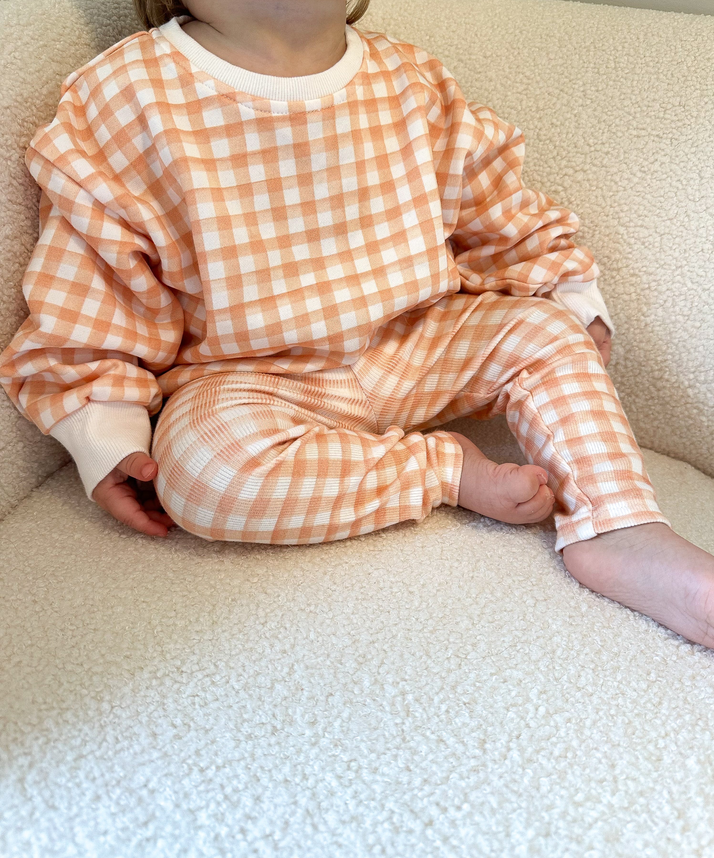 Toddler wearing an orange and white checkered outfit sitting on a beige sherpa chair.