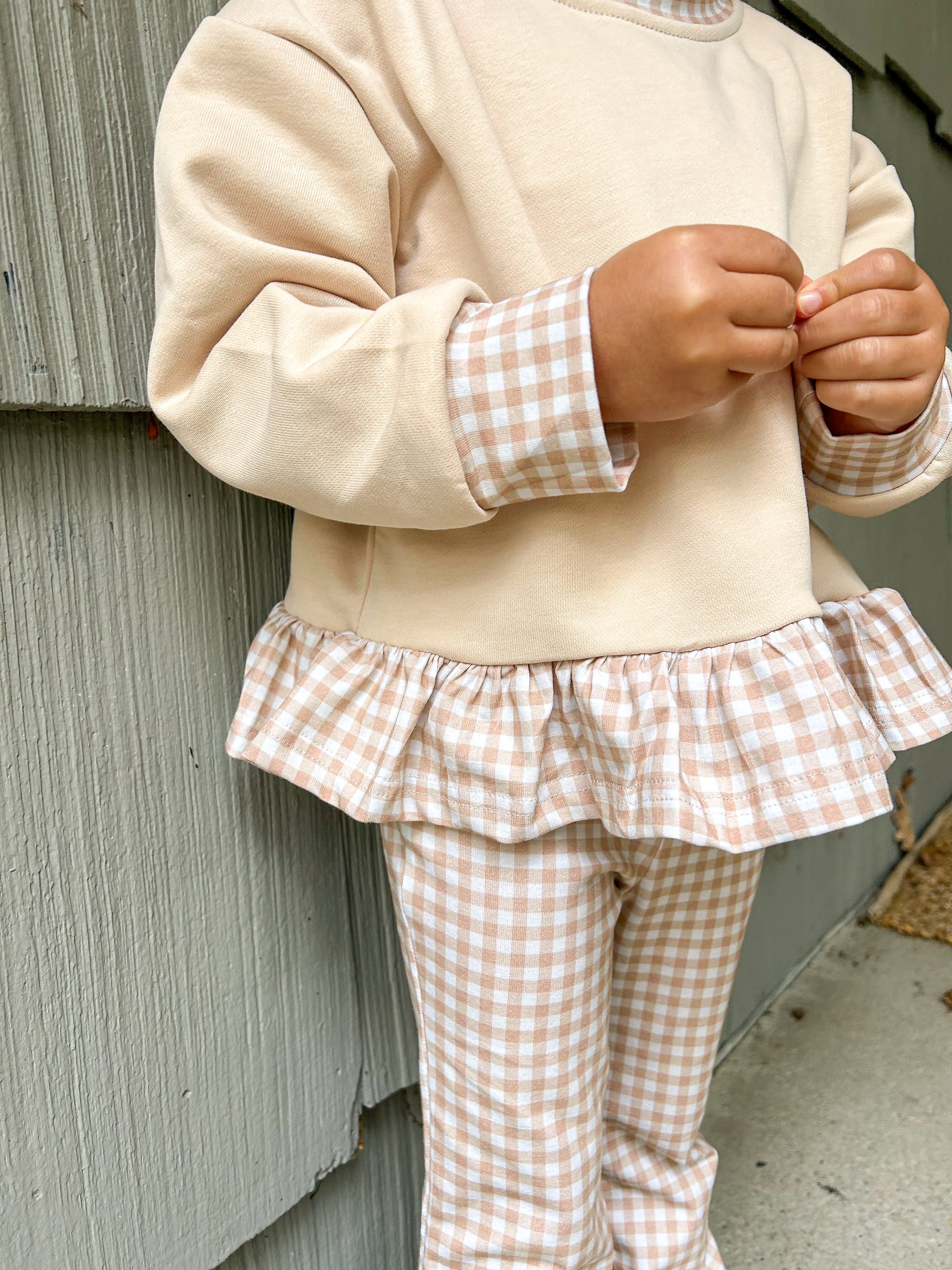 A child wearing a cream sweatshirt with peach-and-white gingham cuffs and a matching ruffled hem, paired with matching gingham pants, standing against a gray wooden wall.