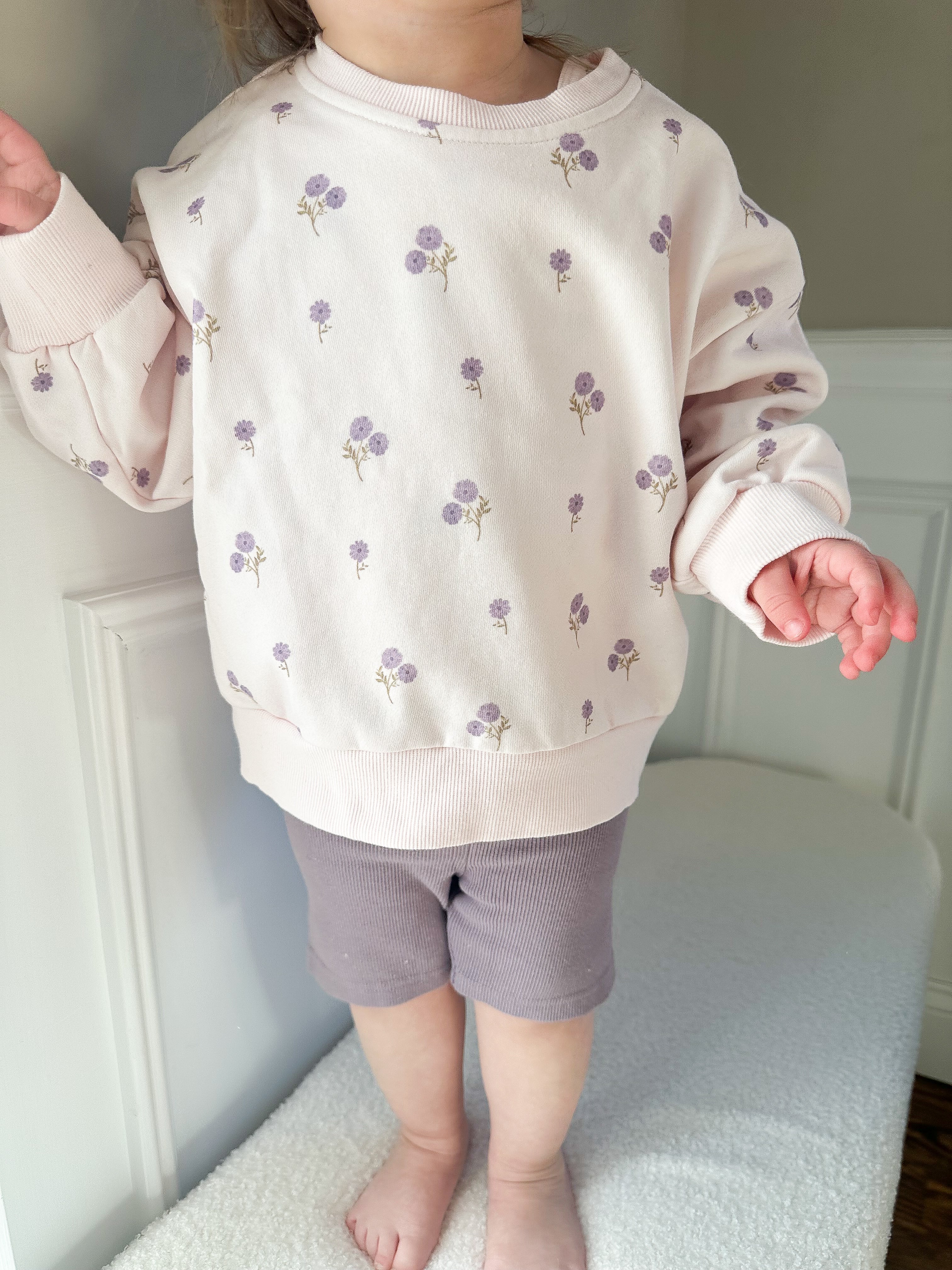 Child wearing a light-colored purple sweatshirt with purple floral patterns and purple biker shorts standing on a beige surface.