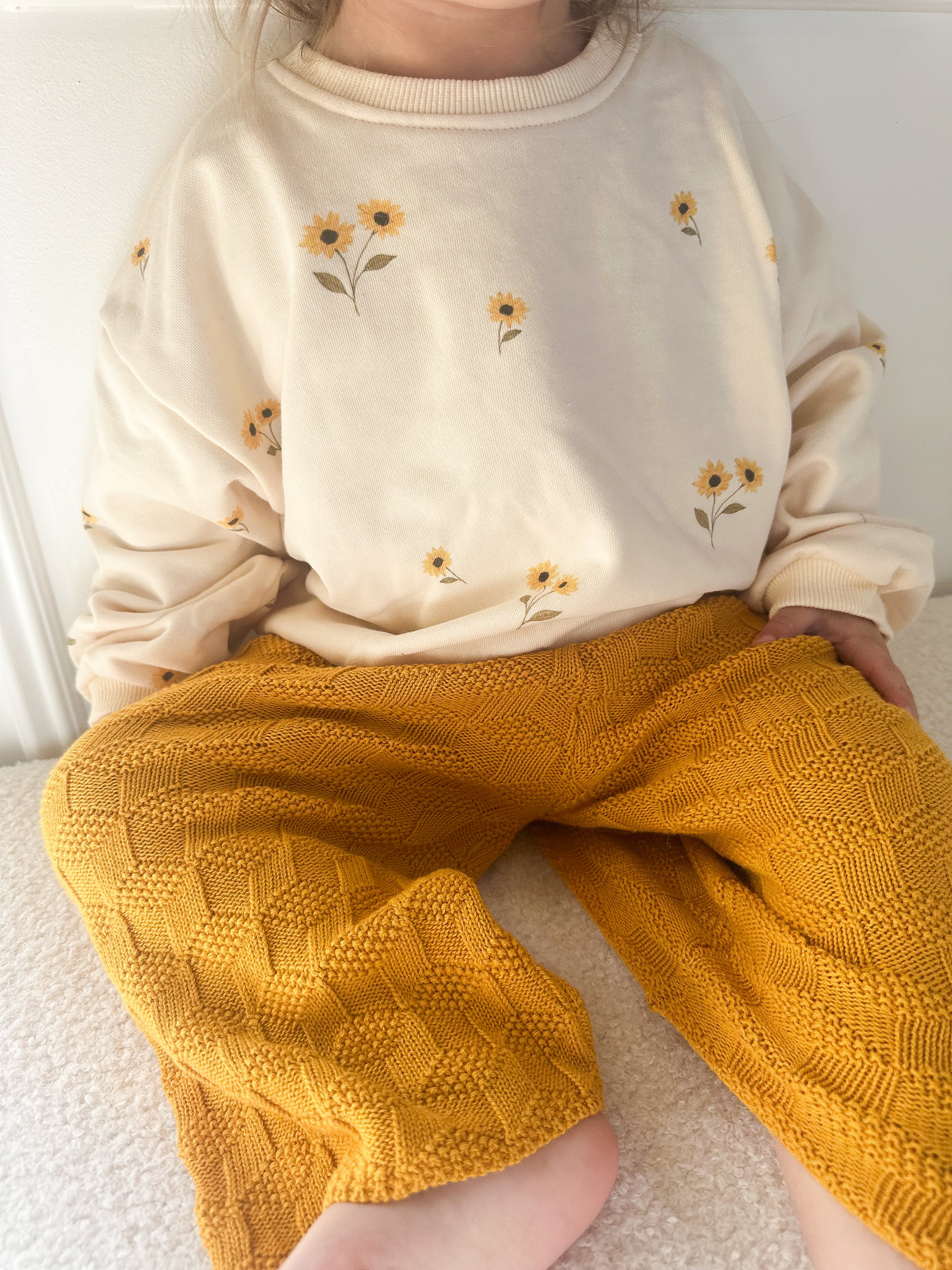 A child wearing a cream sweatshirt with small yellow sunflower prints and mustard yellow textured knit pants, sitting indoors on a light surface.