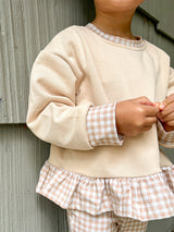 A child wearing a cream sweatshirt with peach and white gingham trim, collar, cuffs, and ruffle hem, paired with matching gingham pants, standing against a gray wooden wall.