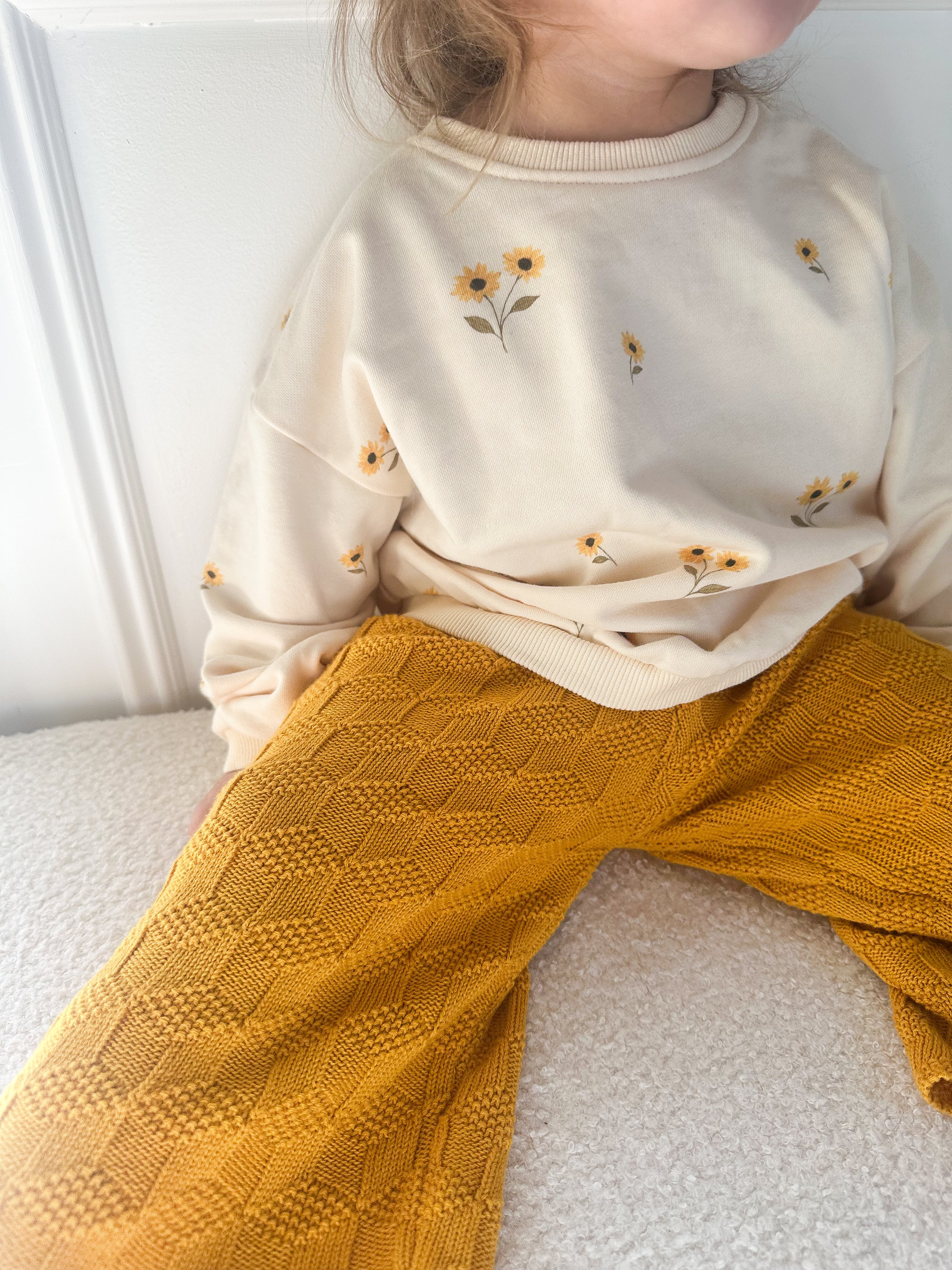 A child wearing a cream sweatshirt with small yellow sunflower prints and mustard yellow textured knit pants, sitting indoors against a white wall.