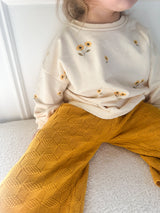 A child wearing a cream sweatshirt with small yellow sunflower prints and mustard yellow textured knit pants, sitting indoors against a white wall.