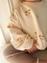 A close-up of a child wearing a cream-colored sweatshirt with yellow sunflowers and brown ribbed pants.