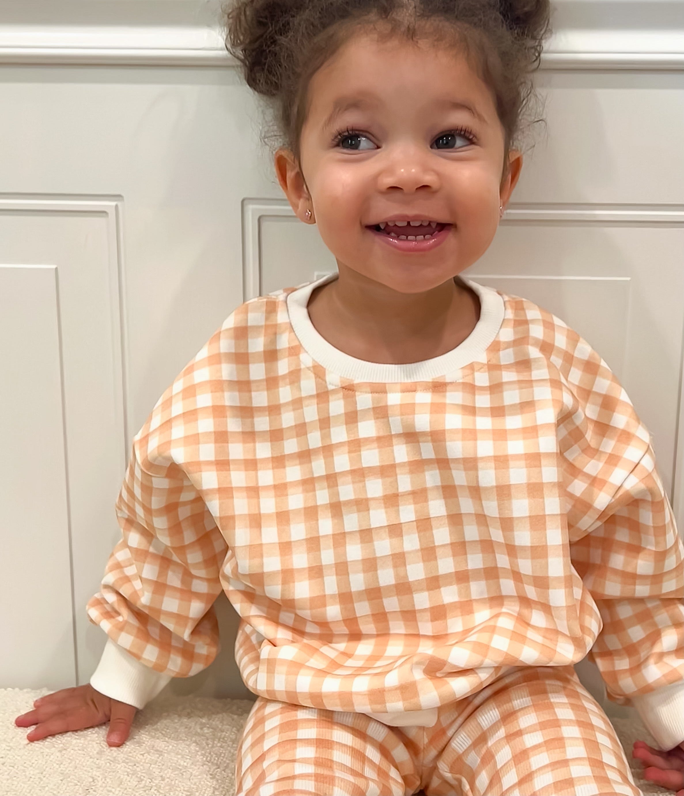 Toddler wearing an orange and white checkered outfit sitting on a beige sherpa ottoman.