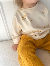 A child wearing a cream sweatshirt with small yellow sunflower prints and mustard yellow textured knit pants, sitting indoors on a light cream surface.