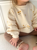 A child wearing a cream sweatshirt with small yellow and green sunflower prints, paired with brown pants, sitting on a beige surface.