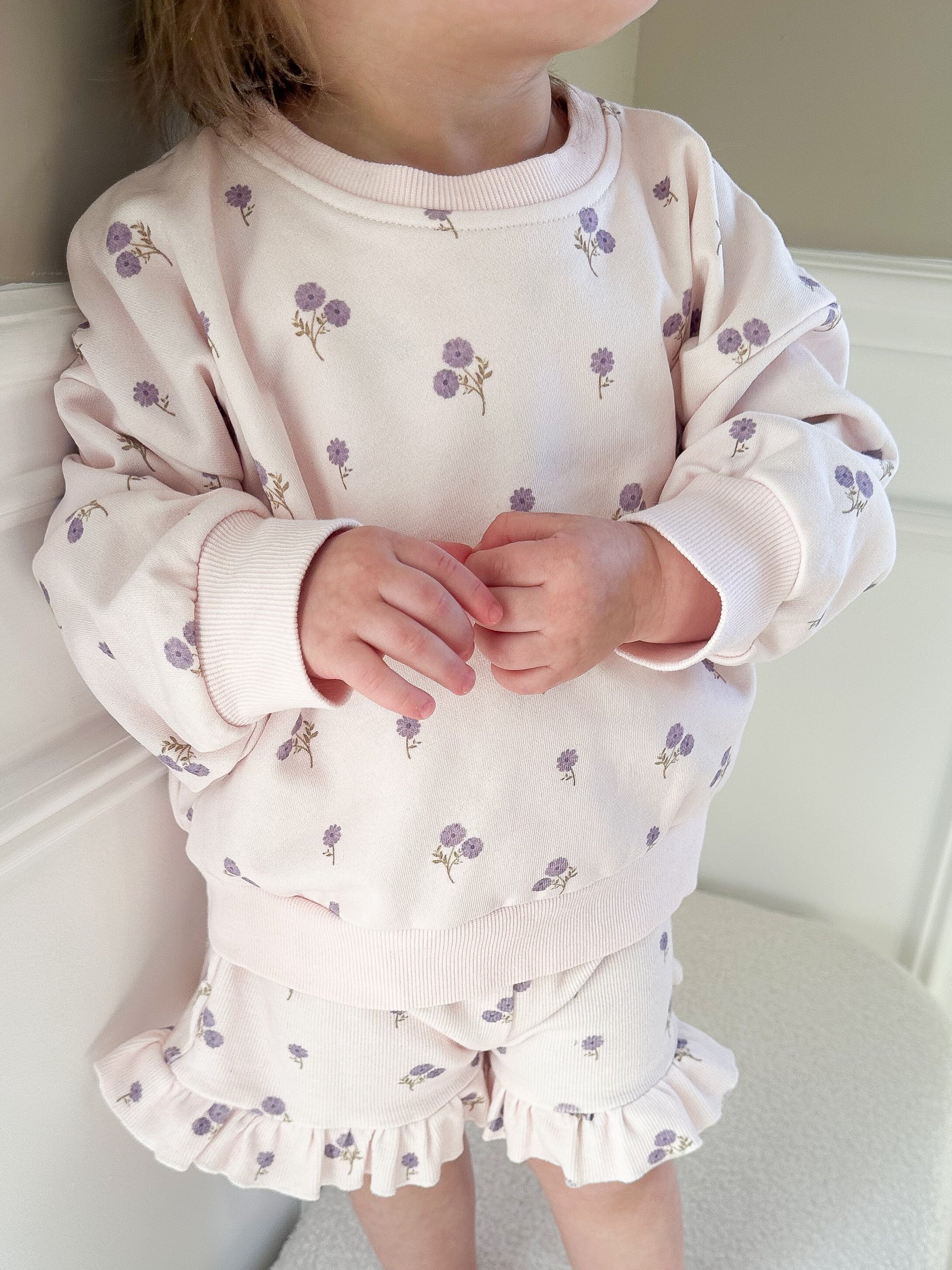 Child wearing a light purple long sleeve outfit and ruffle shorts set standing on a beige surface.