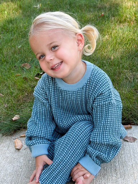 A young child with blonde hair sitting outdoors on a concrete surface with grass in the background, wearing a matching blue and white waffle-knit long-sleeve top and pants set.