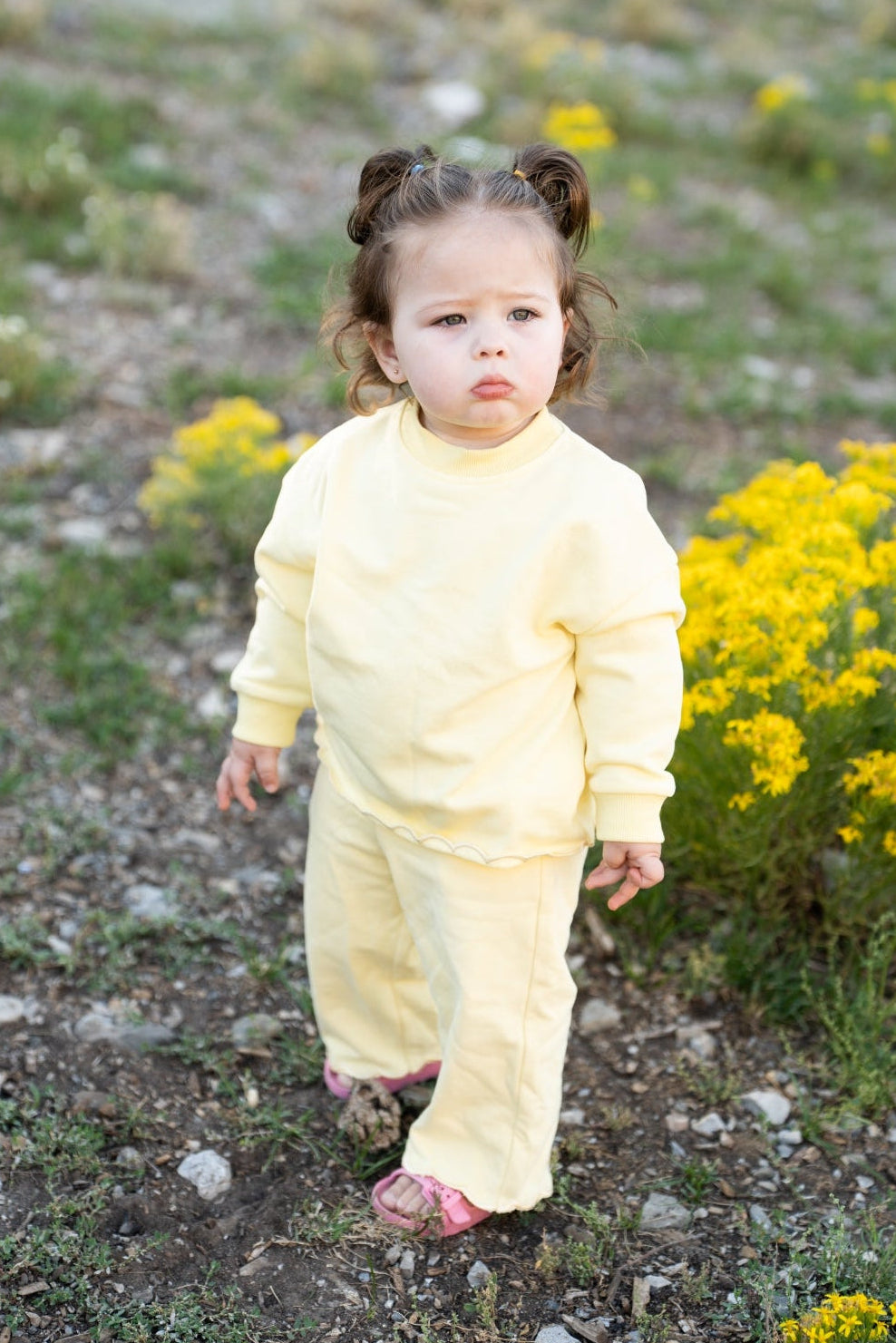 A toddler girl wearing a pale yellow long-sleeve top and matching pants walks outdoors on a rocky path with yellow wildflowers in the background.