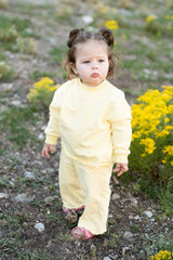 A toddler girl wearing a pale yellow long-sleeve top and matching pants walks outdoors on a rocky path with yellow wildflowers in the background.