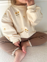 A child sitting indoors wearing a cream sweatshirt with small yellow sunflower prints and brown ribbed pants.