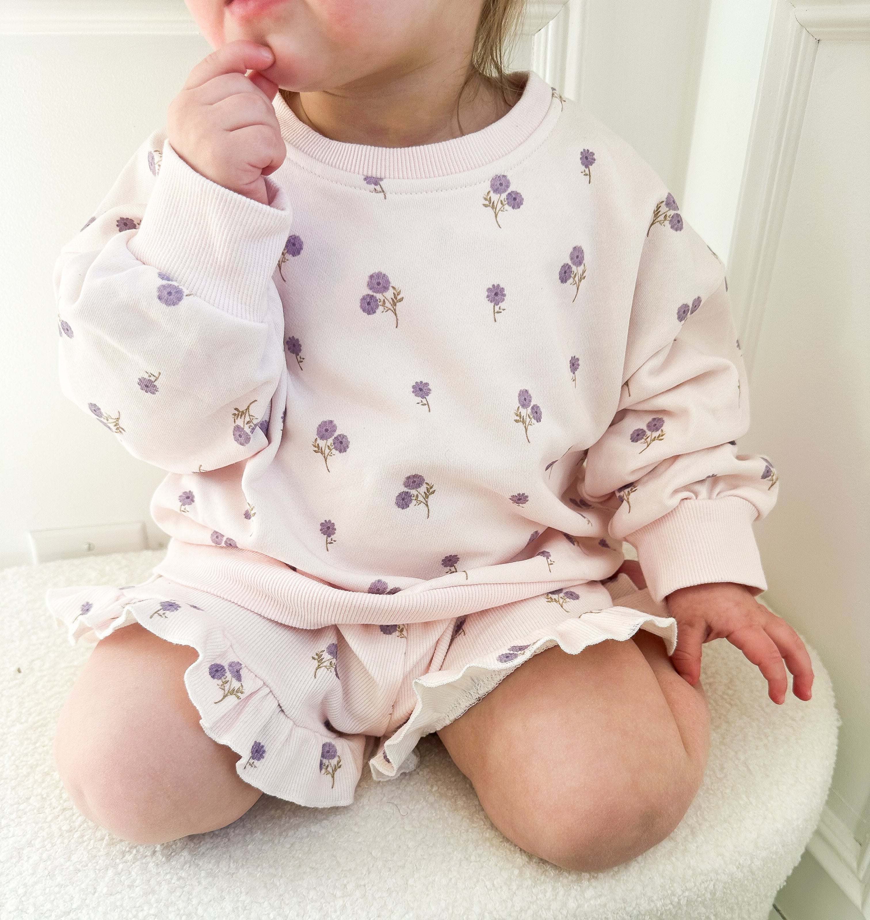 Child wearing a light purple floral outfit sitting on a light beige surface.