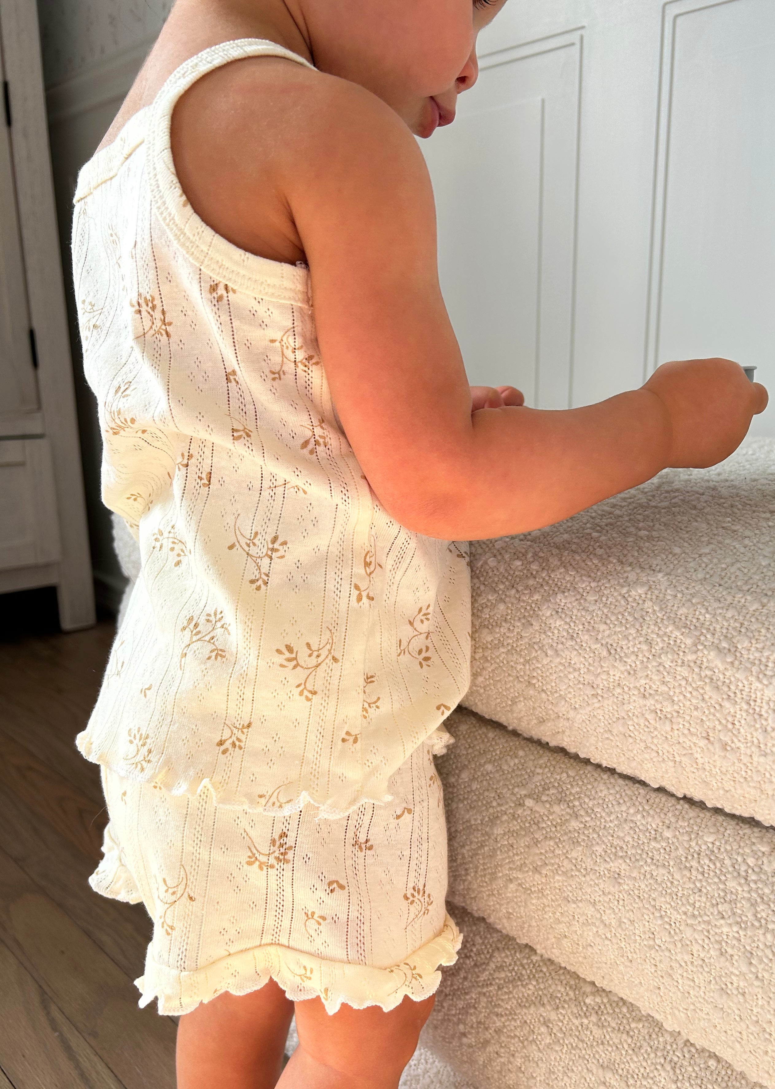 Baby in a white floral tank top and shorts set with ruffles dress standing against bench.