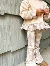 A child wearing a cream sweatshirt with gingham checked cuffs and a ruffled hem, paired with matching gingham pants, standing against a gray wooden siding wall.