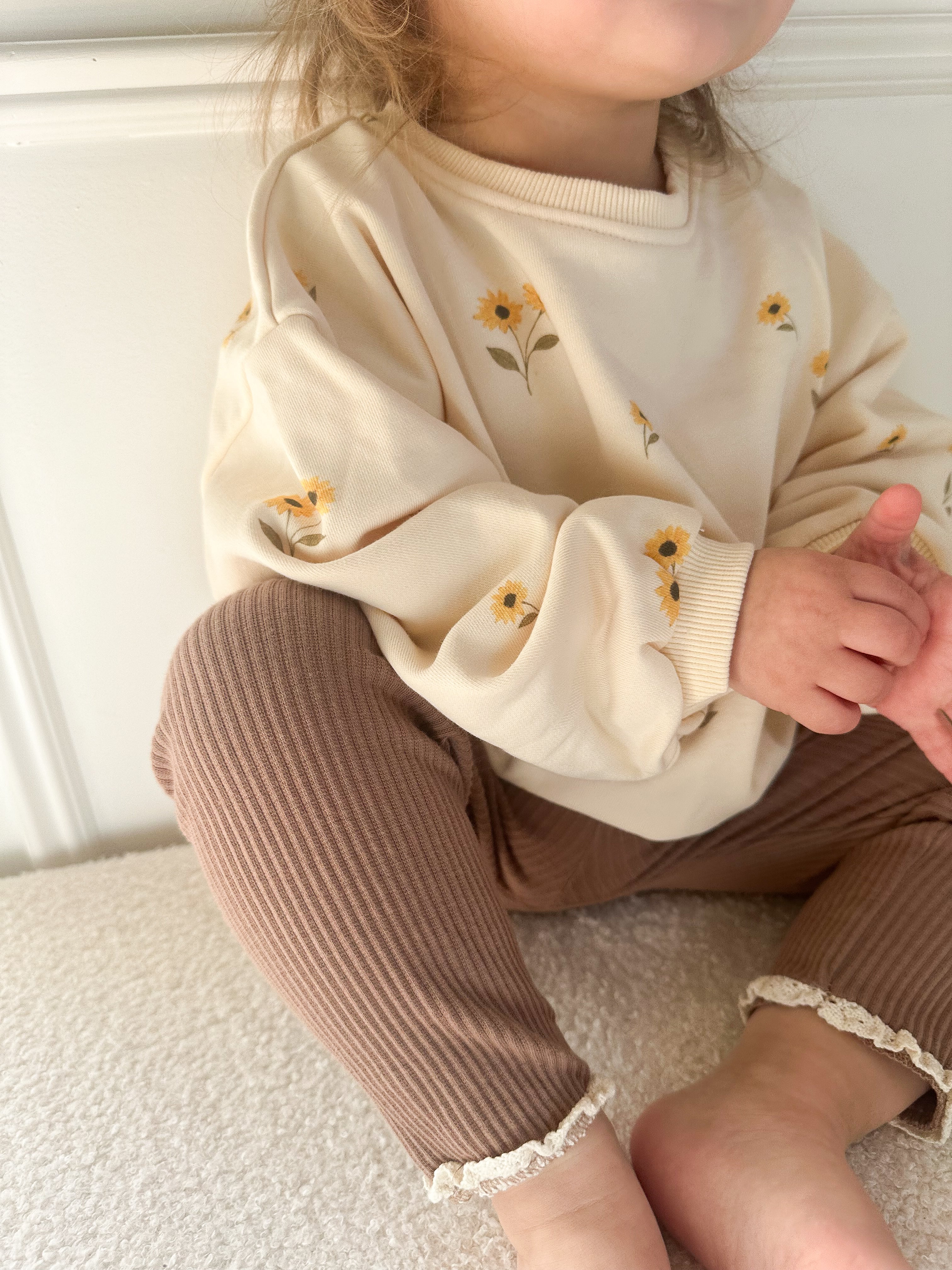 A child wearing a cream sweatshirt with yellow sunflower prints and brown ribbed pants.