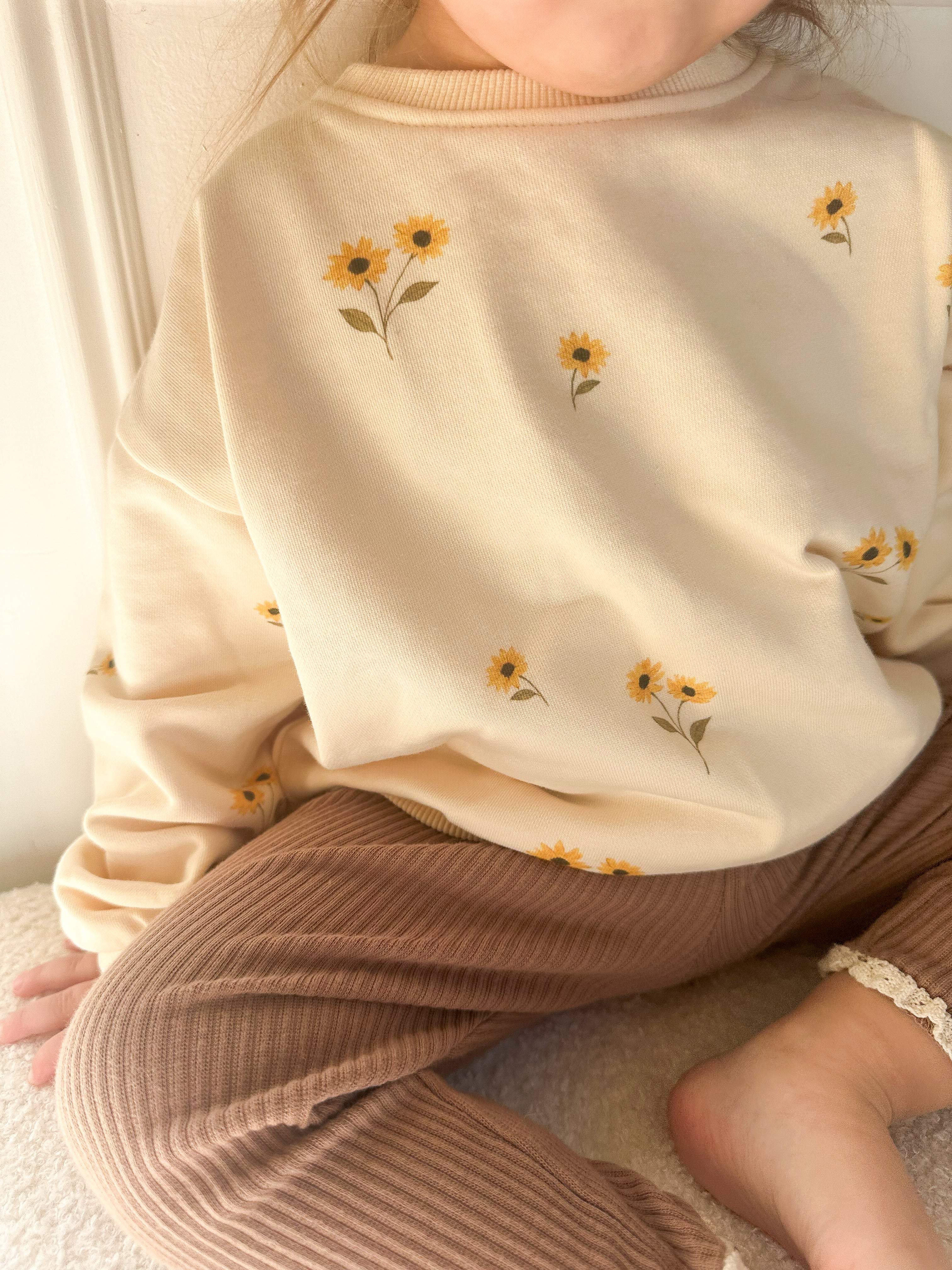 A close-up of a child wearing a cream sweatshirt with yellow sunflower prints and brown ribbed pants, sitting indoors.