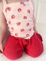 Child wearing a pink outfit with strawberry pattern and red knitted pants on a white background.