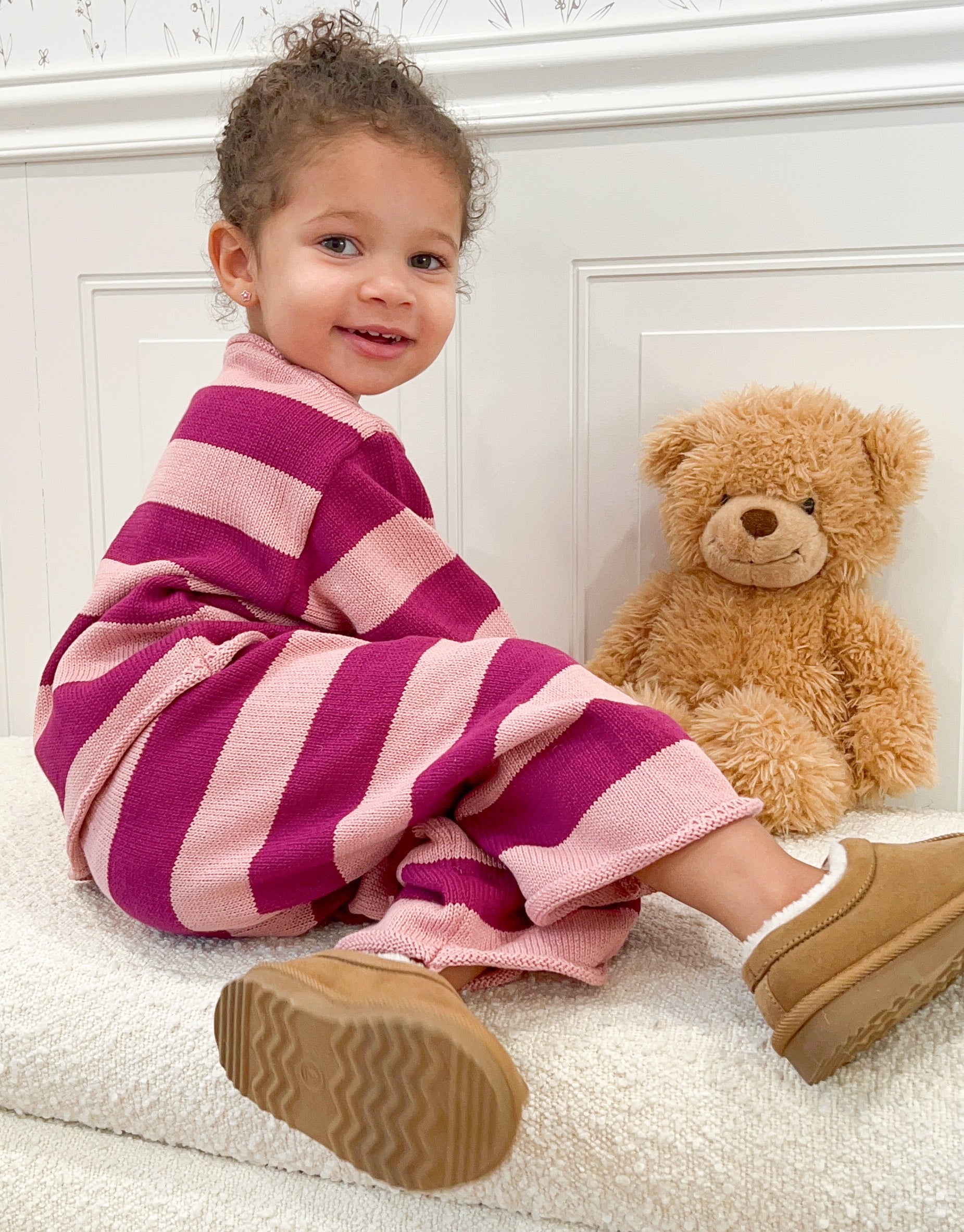 Toddler in pink and purple striped outfit with teddy bear on white background.