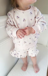 Child wearing a light purple floral long sleeve and ruffle shorts outfit set standing on light-colored surface.