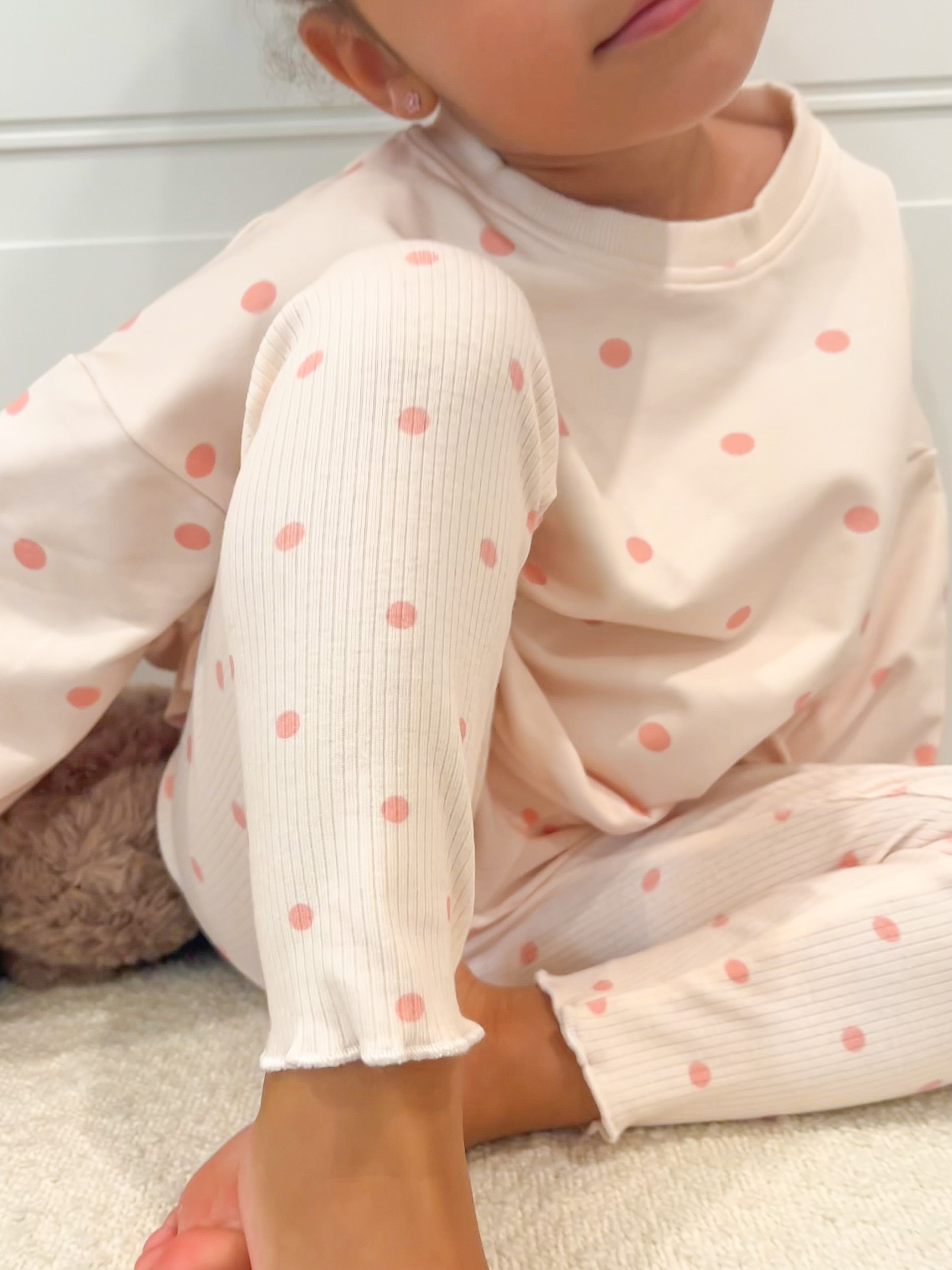 A child wearing a cream-colored ribbed long-sleeve top and matching pants with pink polka dots, sitting on a light carpeted floor.
