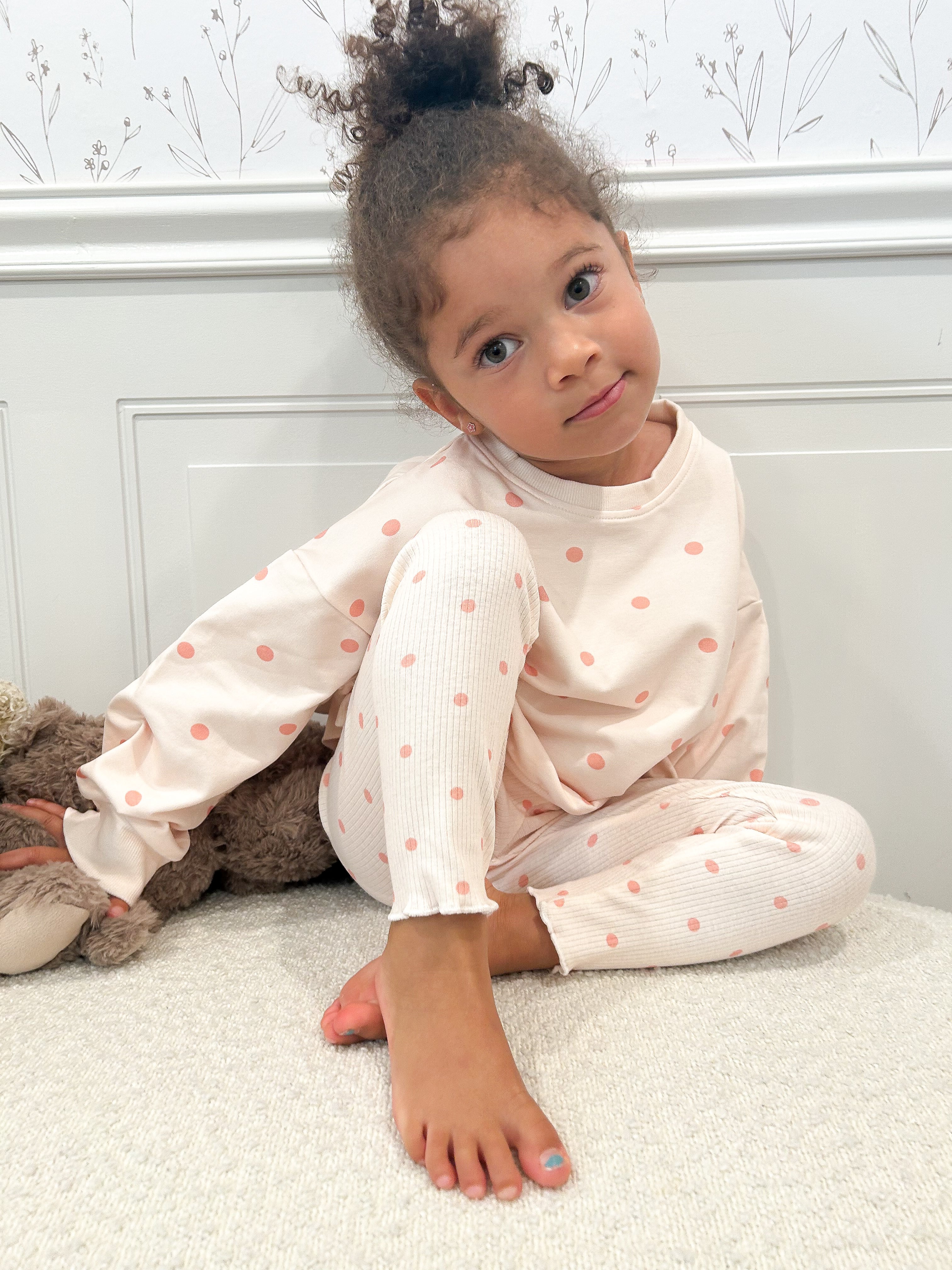 A young child with curly hair sitting on a white carpet in front of a white paneled wall, wearing a light pink long-sleeve top and matching pants with small peach polka dots.