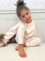 A young child with curly hair sitting on a white carpet in front of a white paneled wall, wearing a light pink long-sleeve top and matching pants with small peach polka dots.