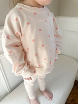 A child wearing a cream-colored sweatshirt and matching pants set with pink polka dots, standing barefoot on a white textured bench in an indoor setting.