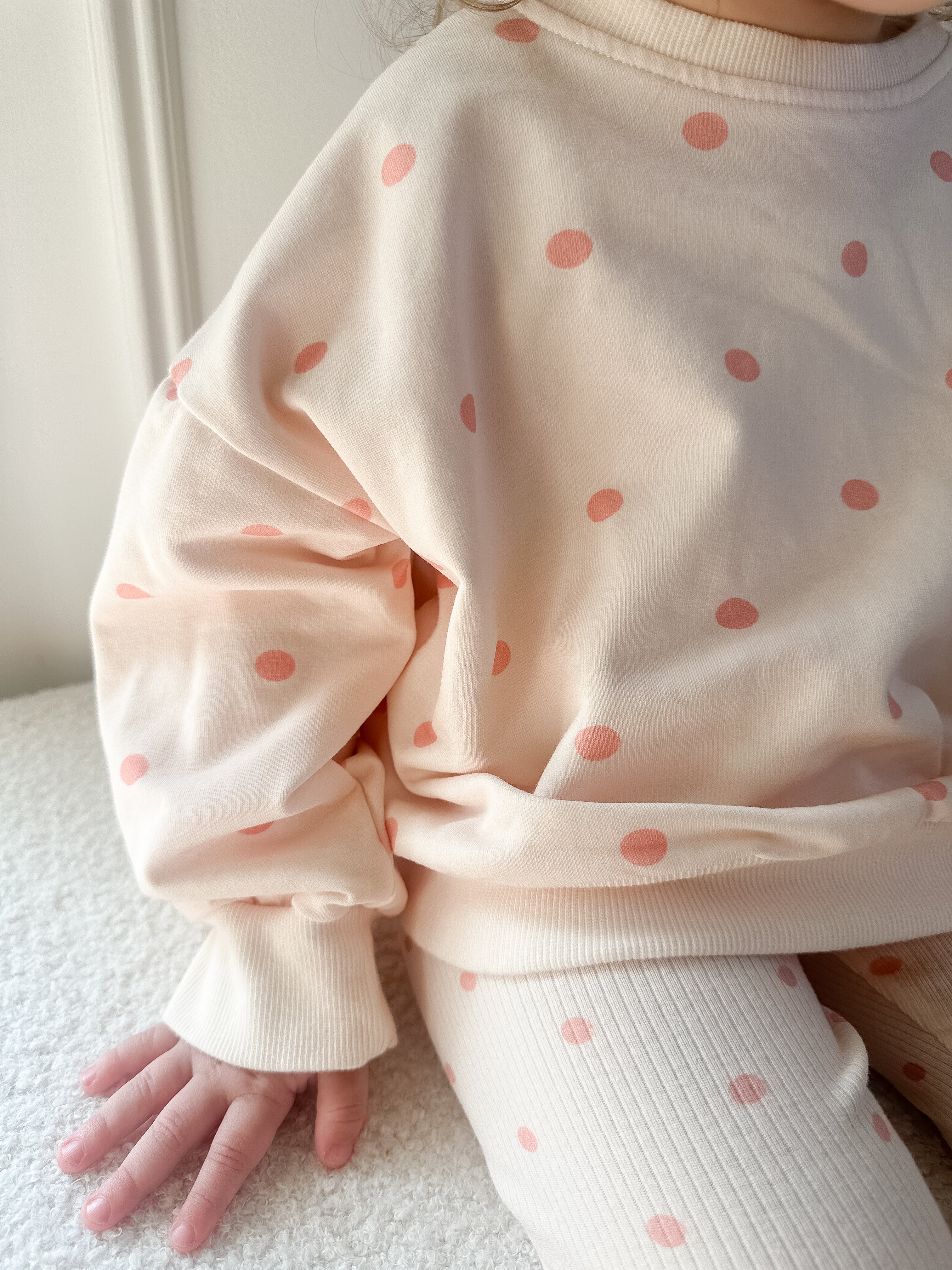 A close-up of a child wearing a cream sweatshirt and matching ribbed leggings with pink polka dots, sitting on a white textured surface.