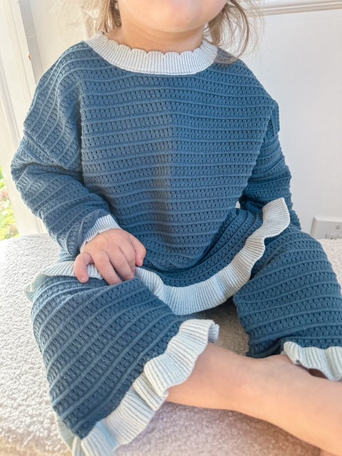 A child wearing a textured blue knit set with light blue ruffled trim, sitting on a textured surface.