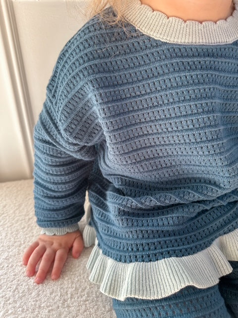A close-up of a child wearing a textured knit sweater in dusty blue with contrasting light blue ruffled trim at the collar and hem, sitting on a light-colored carpet.