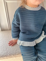 A close-up of a child wearing a textured knit two-piece outfit in dusty blue with contrasting light blue ruffled trim at the collar and hem, sitting on a white textured surface.