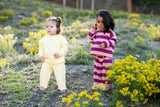 Two young toddlers standing outdoors in a grassy field with yellow flowers. The toddler on the left wears a solid pale yellow long-sleeve top and matching pants.