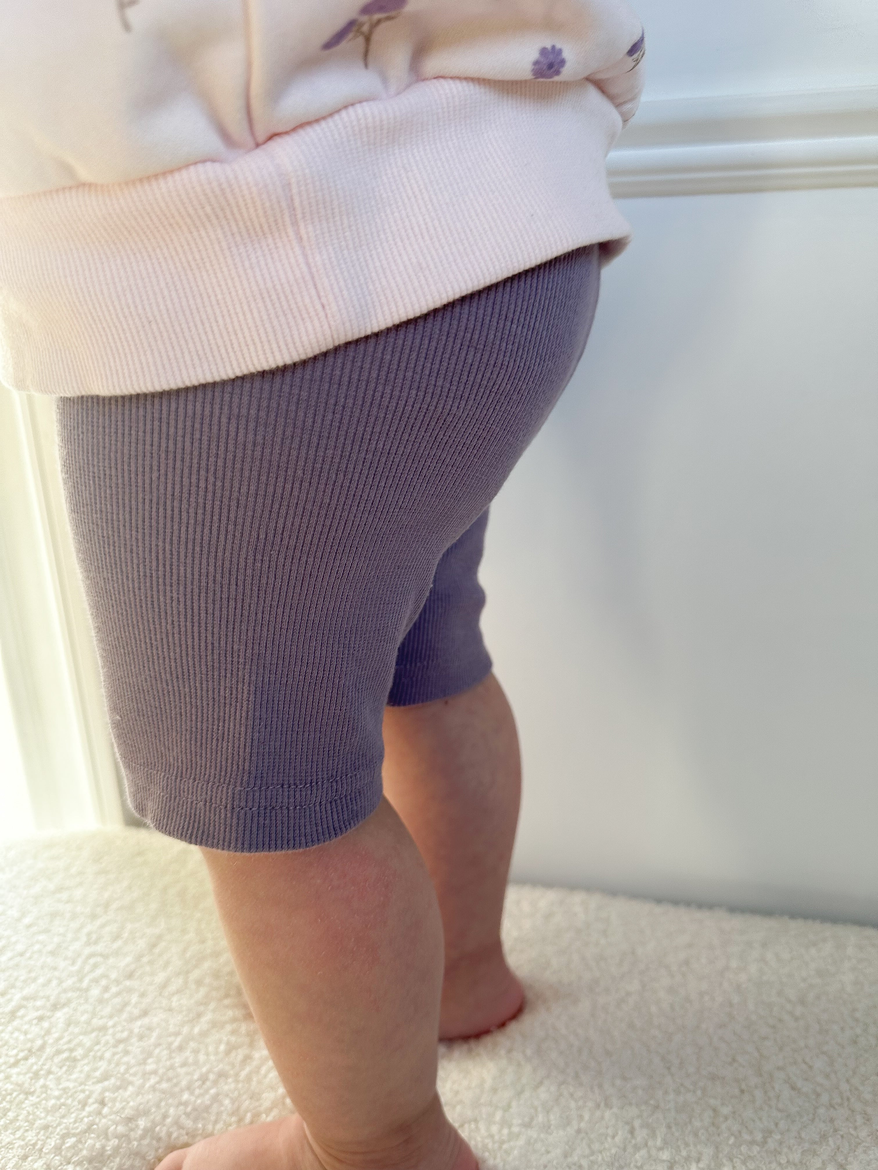 Child wearing purple ribbed biker shorts and a light-colored top standing on a beige surface.